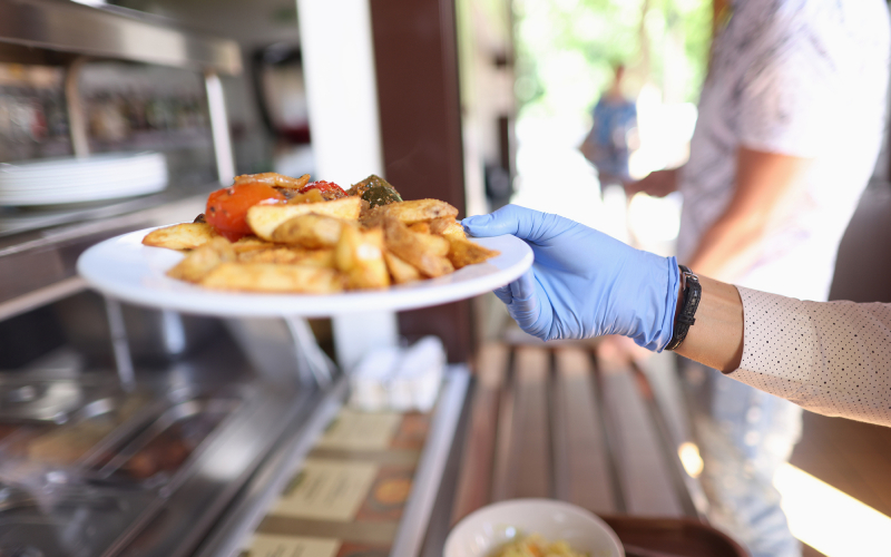 Female hand in blue medical glove hold white plate with fried potatoes and stewed vegetables. Self service restaurant with tray.